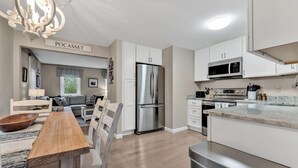 A modern kitchen with white cabinetry, stainless steel appliances, and a dining area with a wooden table and chairs. - Family-Friendly Home Near Beach with Fenced Yard   |   Fire Pit, Three Bedroom Plus Office make for Four Unique Sleeping Spaces, Pocasset Home Perfect for Families, Host up to 7, 24 Hr Hospitality (Bourne)