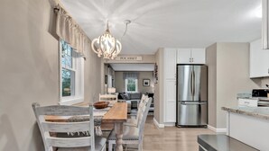 A modern kitchen with a stainless steel refrigerator, a dining area with a wooden table and white chairs, and a chandelier. - Family-Friendly Home Near Beach with Fenced Yard   |   Fire Pit, Three Bedroom Plus Office make for Four Unique Sleeping Spaces, Pocasset Home Perfect for Families, Host up to 7, 24 Hr Hospitality (Bourne)