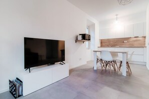 Een moderne leefruimte met een flatscreen televisie bevestigd aan een witte kast, een eethoek met een witte tafel en stoelen, en een keuken met witte kastjes en een houten aanrecht.
