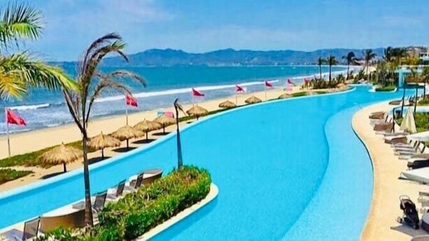 A pool area with a curved design, a beach, and palm trees.