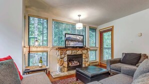 A living room with a stone fireplace, a television, and large windows with blinds.