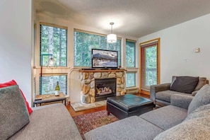 A living room with a stone fireplace, a television, and large windows with blinds. - Greystone Lodge 224 Ski-in/Out by Acer Vacations (Whistler)