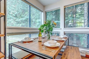 A dining area with a long table set for six, large windows with blinds, and a vase with flowers. - Greystone Lodge 224 Ski-in/Out by Acer Vacations (Whistler)