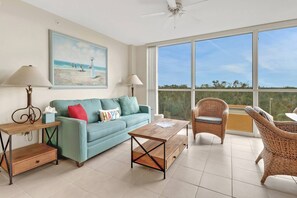 A spacious living room with a teal sofa, a wooden coffee table, wicker chairs, and a large window offering a view of the outdoors.