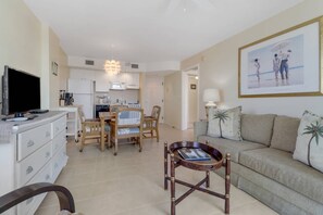 A living room with a TV, a sofa, a coffee table, and a dining area with chairs and a table. - Painted Skies, Estero Bay & Pool Views | Extended Balcony | Pool & Hot Tub | Lovers Key Resort (Fort Myers Beach)