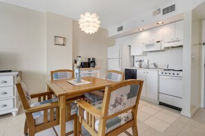 A compact kitchen with white appliances, a dining table with chairs, and a pendant light. - Painted Skies, Estero Bay & Pool Views | Extended Balcony | Pool & Hot Tub | Lovers Key Resort (Fort Myers Beach)