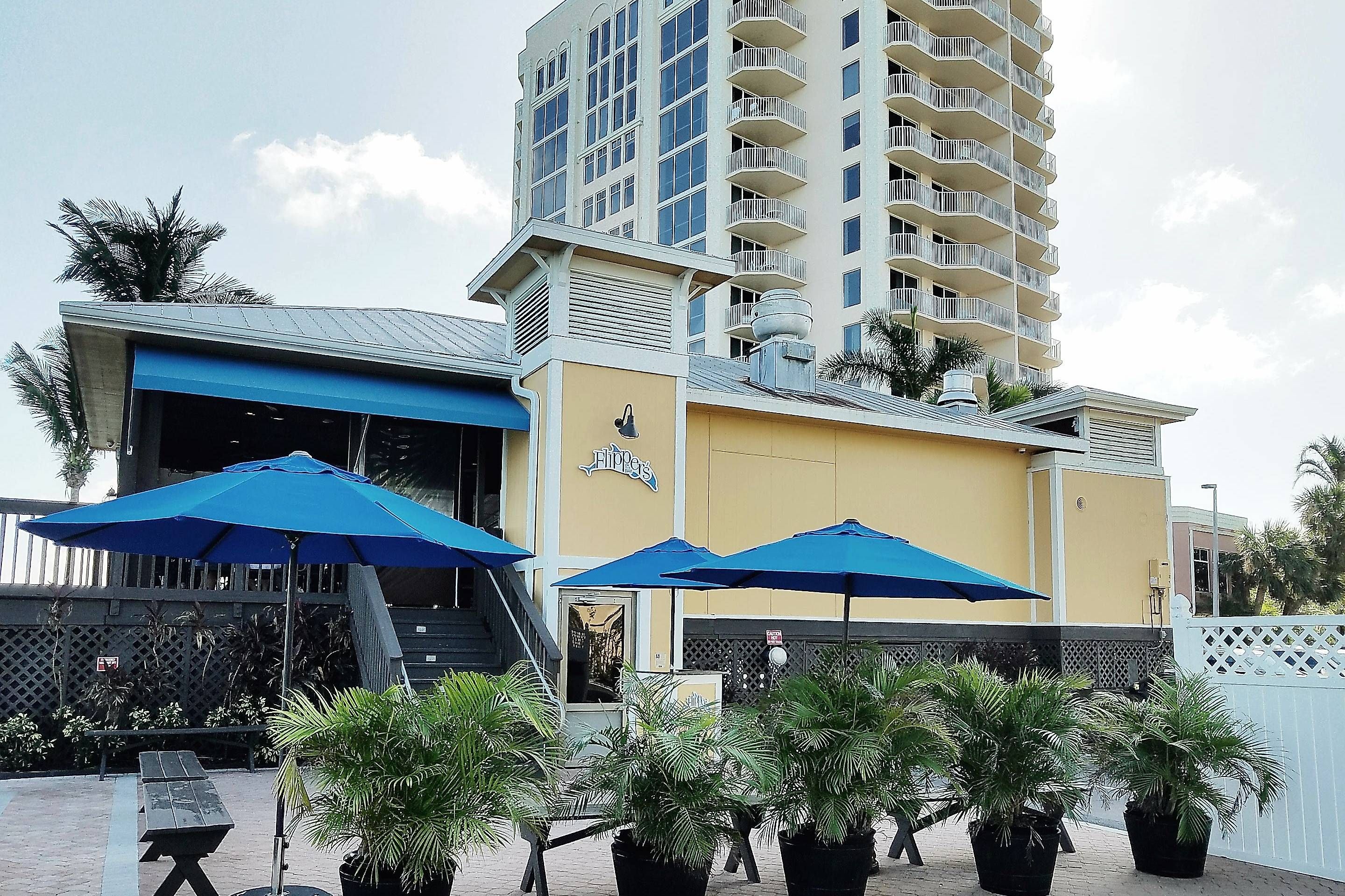 A hotel with a blue awning, outdoor seating with umbrellas, and a tall building in the background.