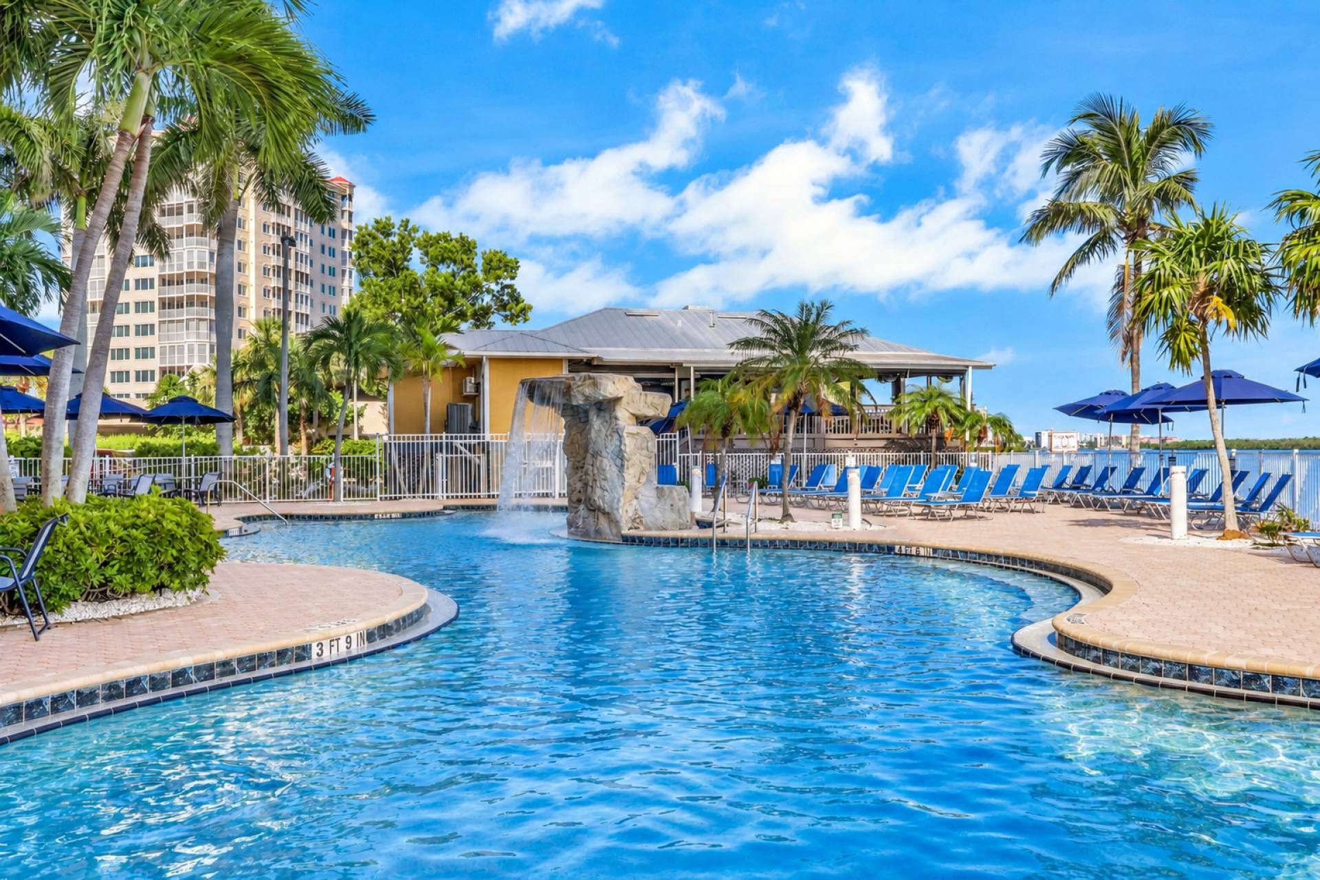 A swimming pool with a waterfall feature, surrounded by lounge chairs and palm trees.