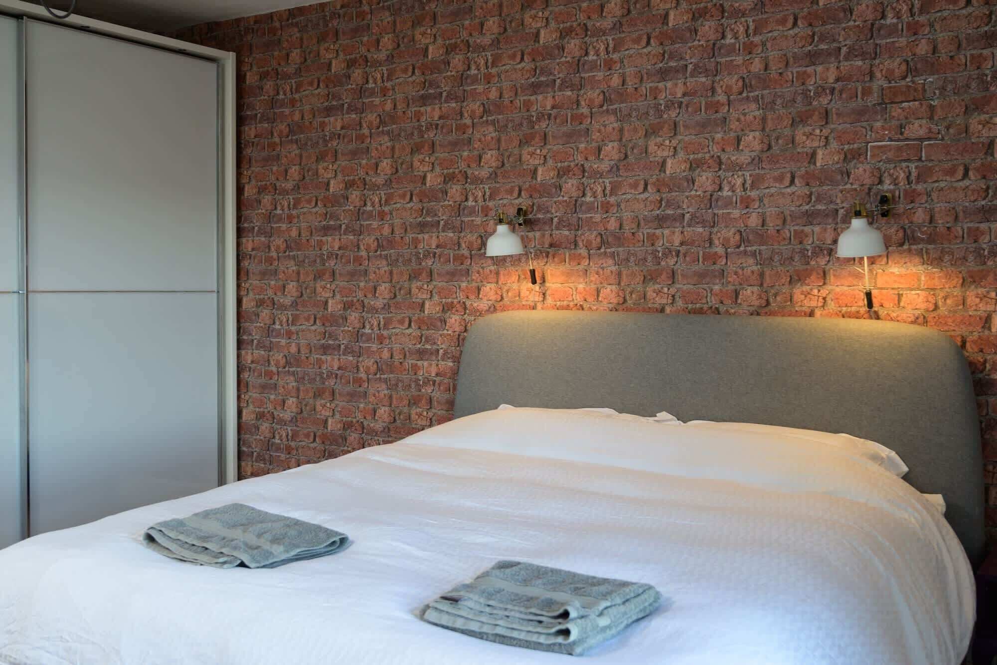 A hotel room with a brick wall, a bed with white bedding, and two wall-mounted lamps.