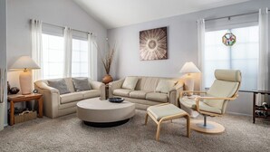 Un salon moderne avec un canapé beige, une table basse ronde et un fauteuil de couleur claire. On aperçoit un vase contenant des plantes séchées sur une table d’appoint, ainsi qu’un grand tableau abstrait accroché au mur.