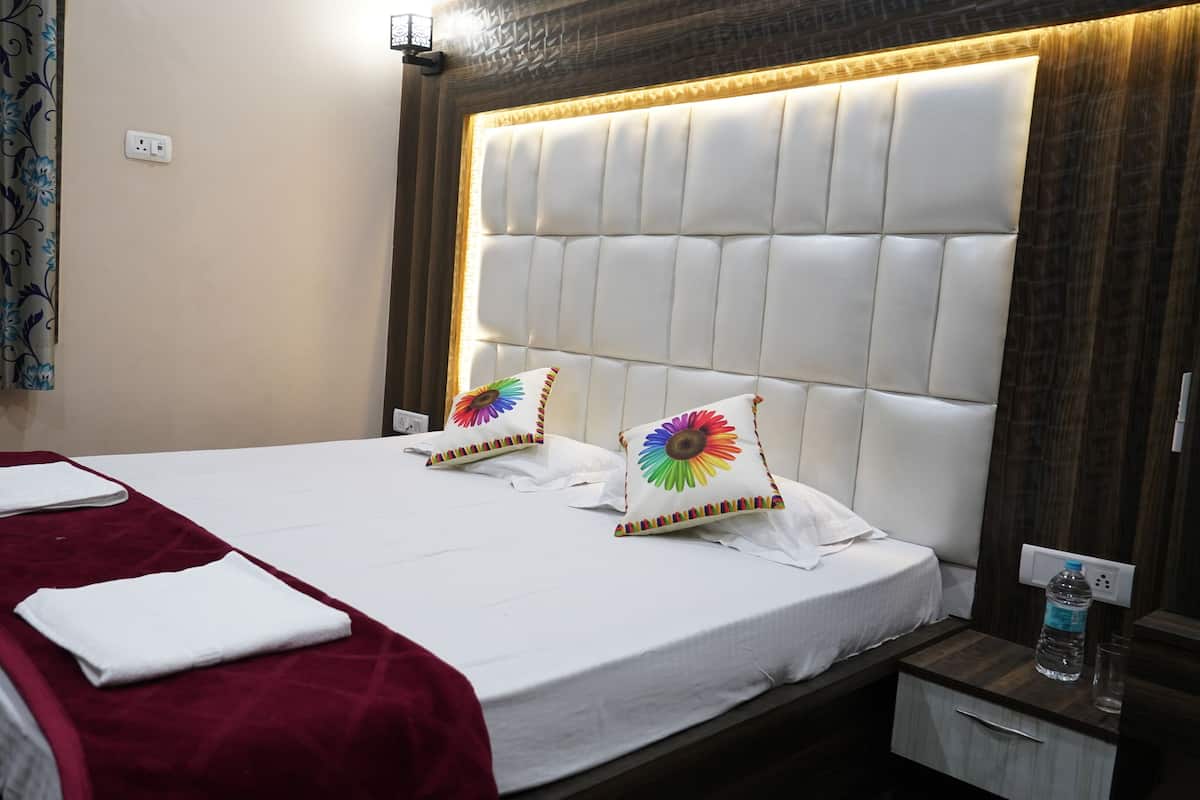 Hotellihuone, jossa on suuri sänky, punainen heittopeitto, kaksi värikkäällä kukkakuviolla koristeltua tyynyä, puinen pääty ja yöpöytä, jossa on vesipullo.