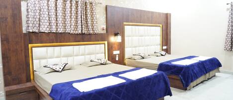 A hotel room with two beds, each with a headboard and a blue bedspread. There are curtains on the windows, and a wooden panel design on the walls.