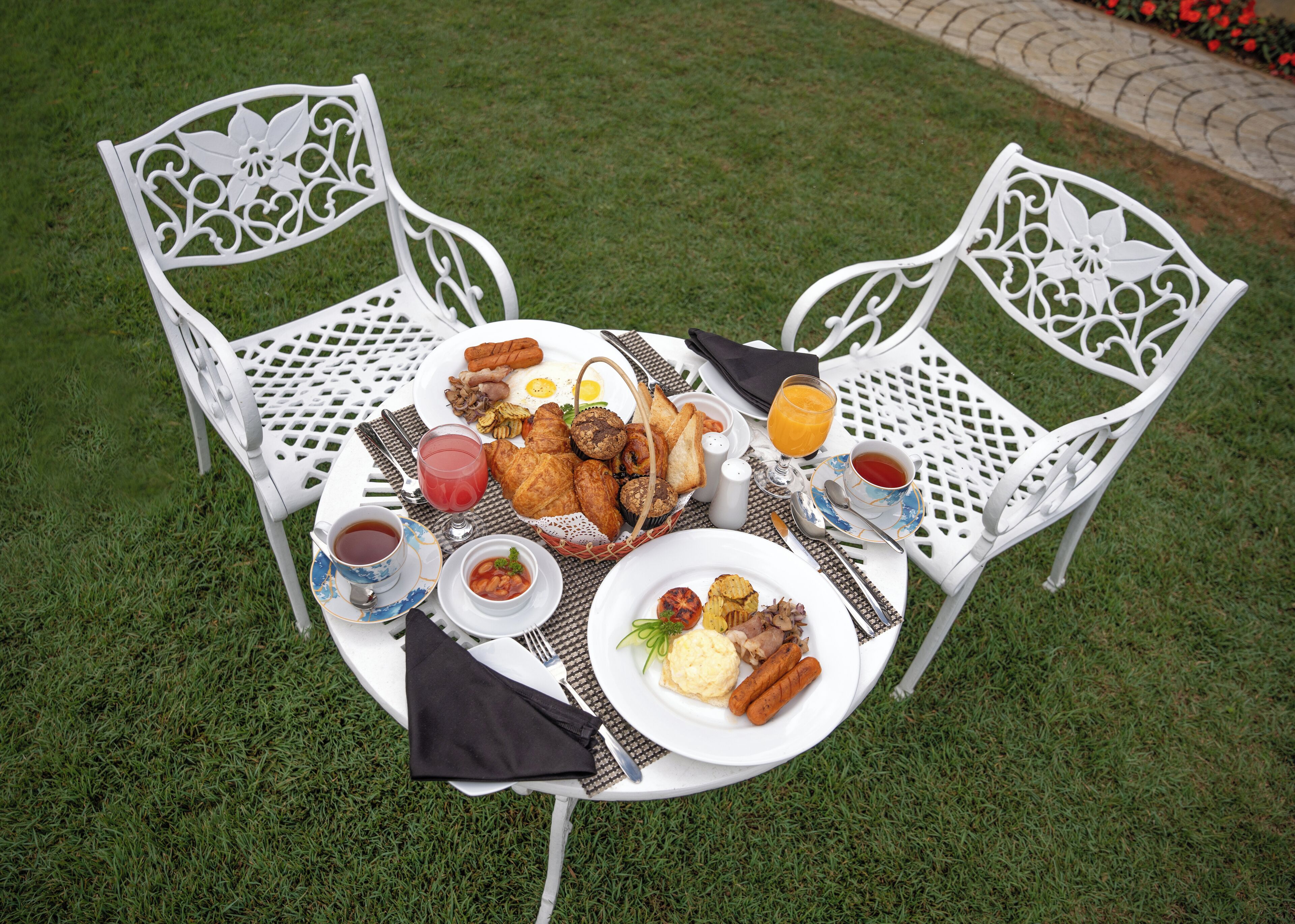 A round table with a white tablecloth, surrounded by white chairs with intricate designs. The table is set with a variety of breakfast items including toast, pastries, eggs, and a hot beverage.