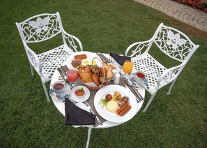 A round table with a white tablecloth, surrounded by white chairs with intricate designs. The table is set with a variety of breakfast items including toast, pastries, eggs, and a hot beverage. - Winchester Little England (Nuwara Eliya)