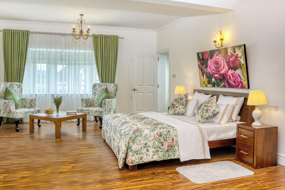 Une chambre à coucher avec une couvre-lit à motifs floraux, deux chaises, une table basse en bois, une table de chevet avec une lampe et un tableau représentant des roses.