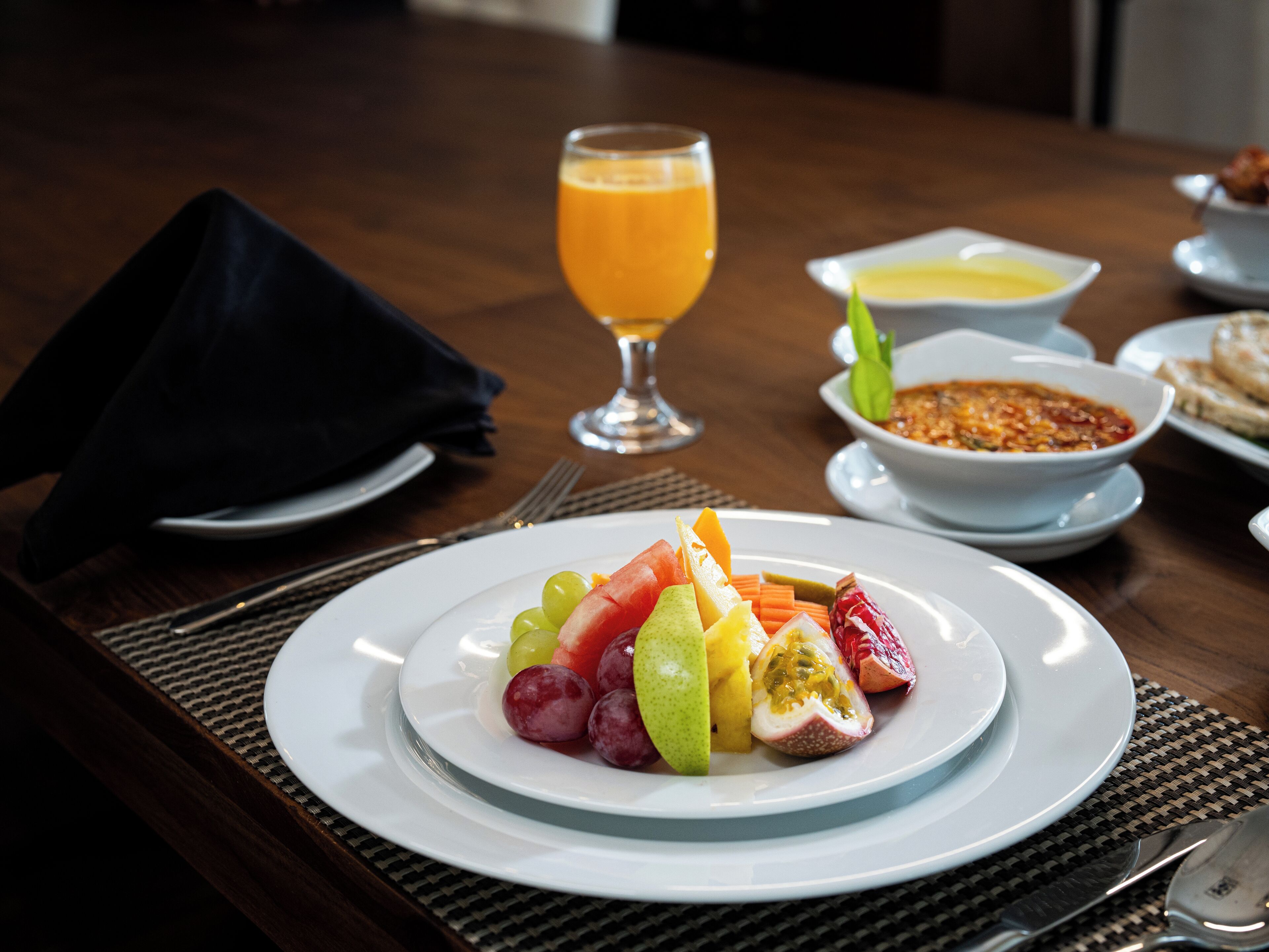 A dining table with a plate of fruit, a glass of orange juice, and a bowl of soup.