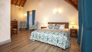 Ein Schlafzimmer mit Holzbalkendecke, einem großen Bett mit floralem Bettbezug, zwei Stühlen, einem Nachttisch mit Lampe und einer Wandleuchte.