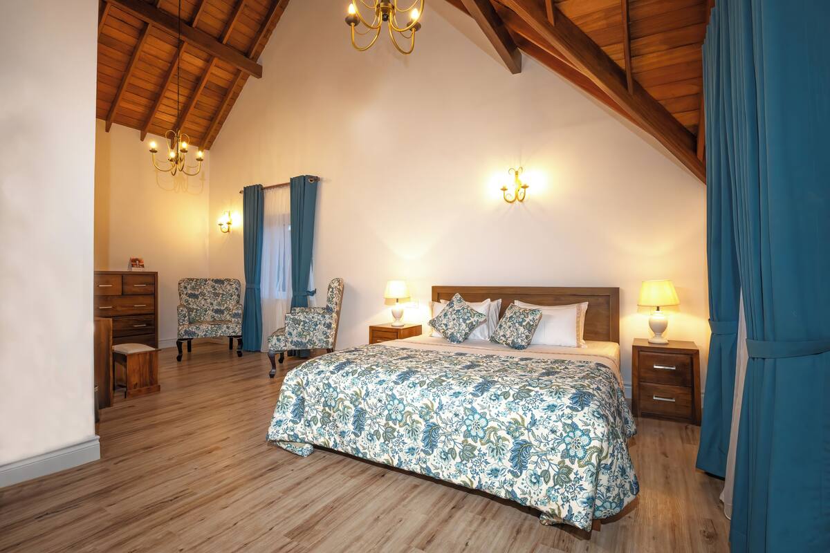 Une chambre avec un plafond en bois, un grand lit avec une literie à motifs floraux, deux chaises, une table de chevet avec une lampe et un luminaire fixé au mur.