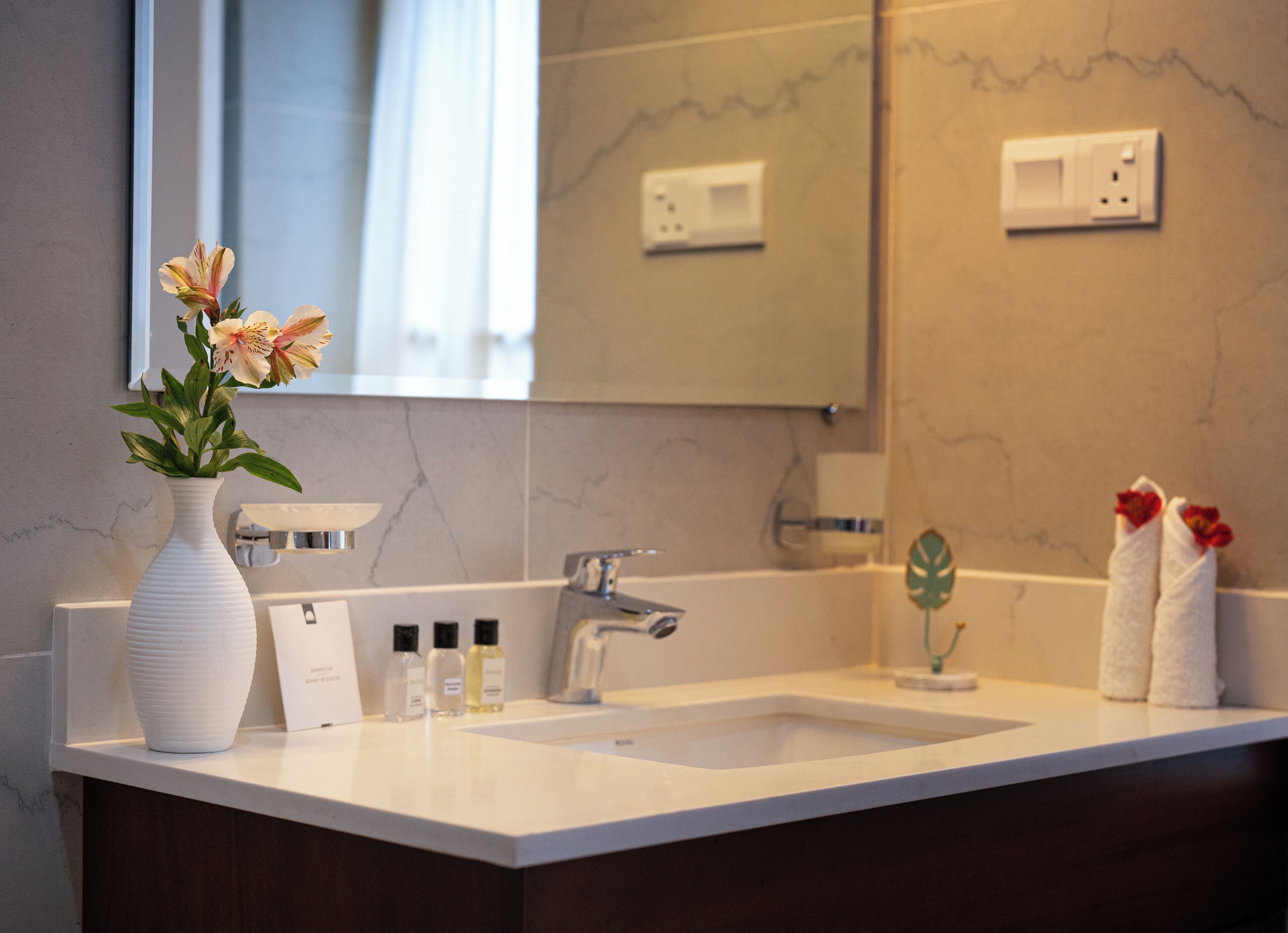 A modern bathroom with a white sink, a mirror, and a vase with flowers. There are toiletries on the counter and towels hanging on the wall.