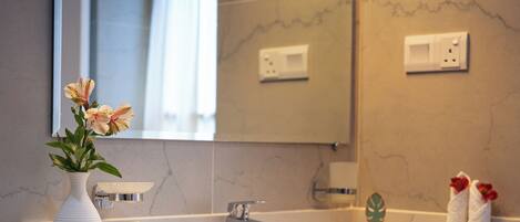 A modern bathroom with a white sink, a mirror, and a vase with flowers. There are toiletries on the counter and towels hanging on the wall.