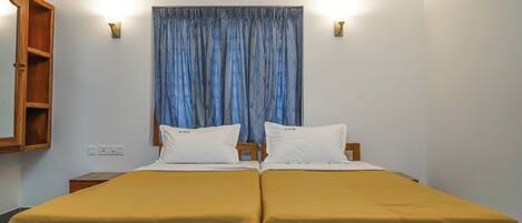 A double bed with a wooden frame, two white pillows, and a yellow bedspread. There is a wooden nightstand to the left, a wall-mounted light, and a ceiling-mounted air conditioner.