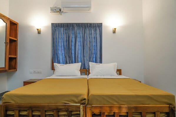 A double bed with a wooden frame, two white pillows, and a yellow bedspread. There is a wooden nightstand to the left, a wall-mounted light, and a ceiling-mounted air conditioner. - Aloha Homestay (Varkala)
