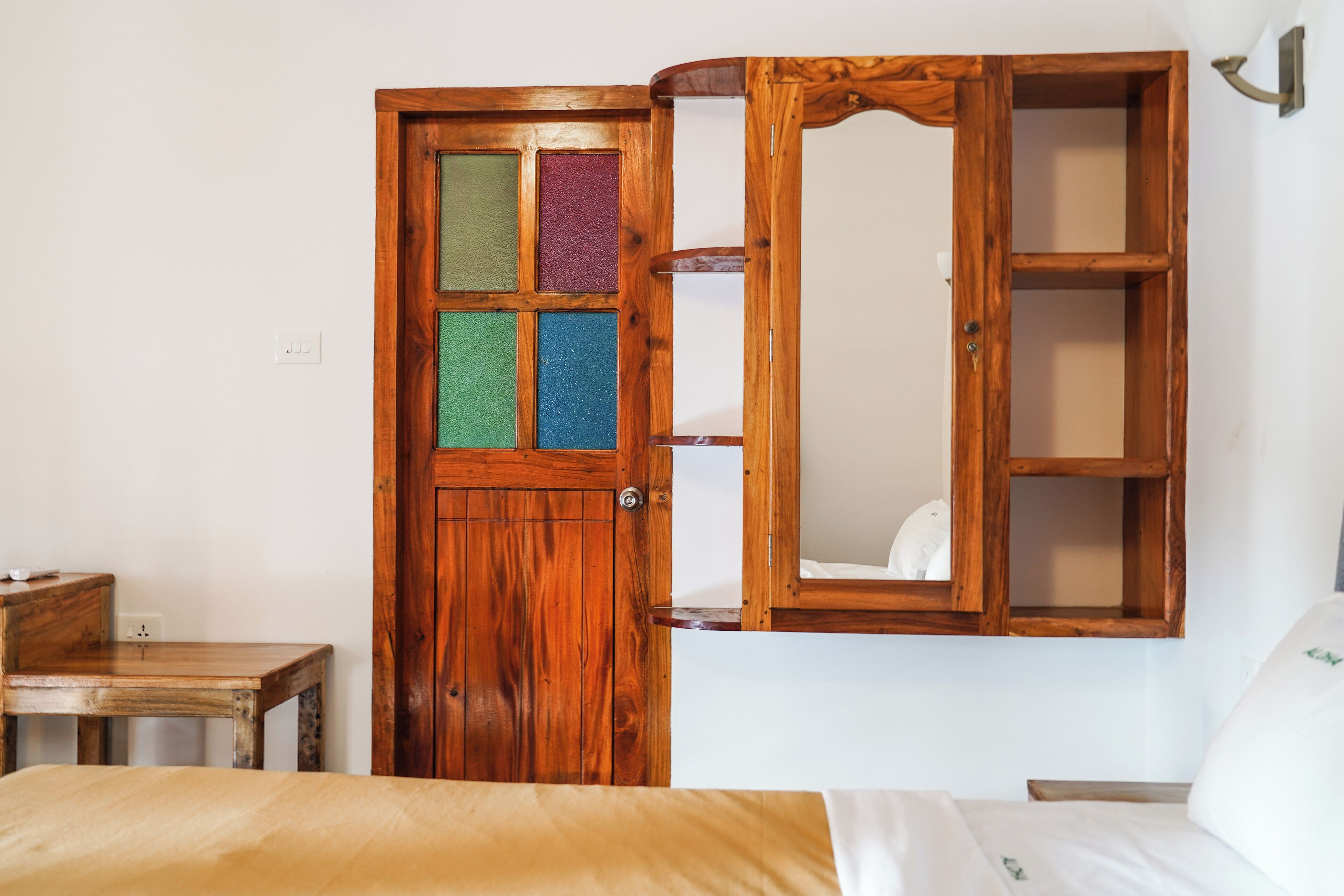 Una porta in legno con uno specchio e una mensola, un letto con materasso bianco e un piccolo tavolino in legno.