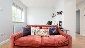 A modern living room with a red velvet sofa, a radiator, a window, and a bookshelf. - Stylish Brixton Hill Studio with Modern Charm (London)