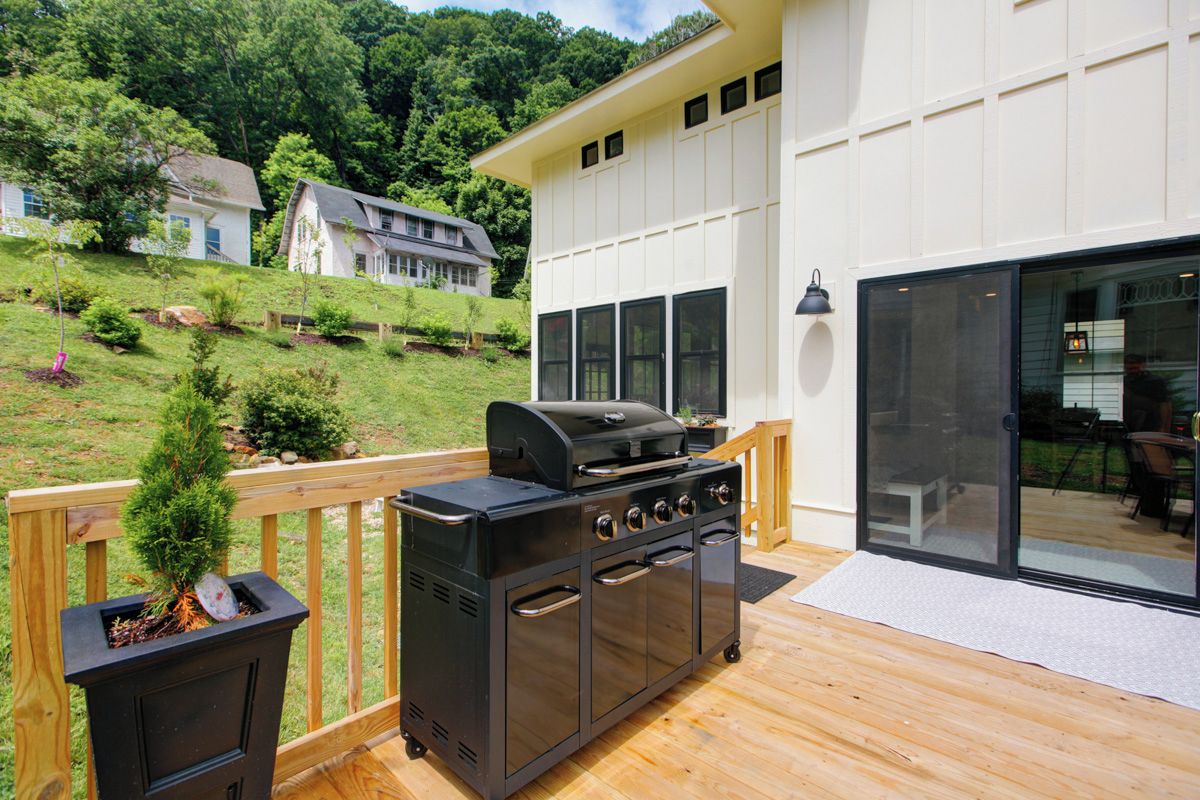A deck with a barbecue grill, a planter with a small tree, and a view of a house in the background.