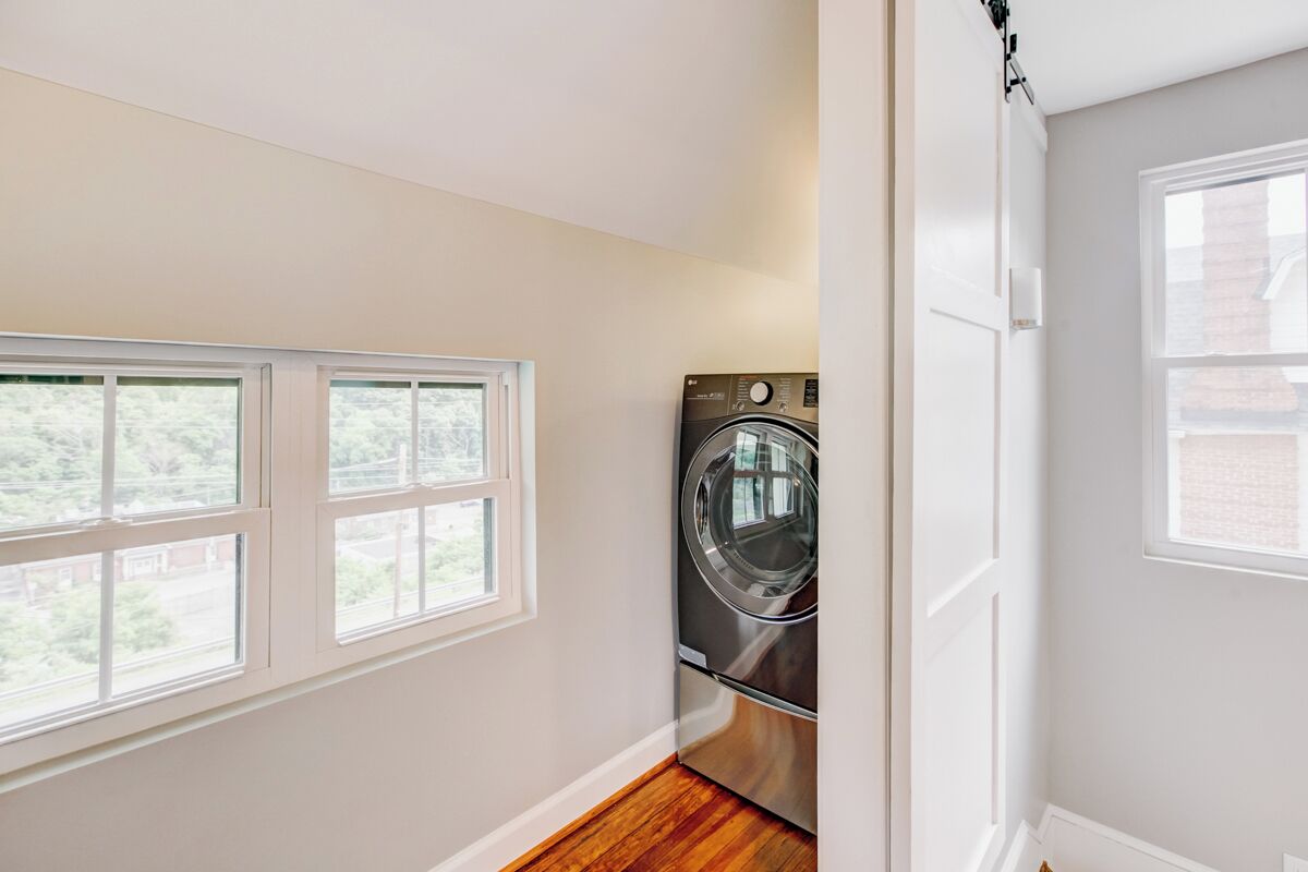 A laundry room with a front-loading washing machine, large windows, and a wooden floor.