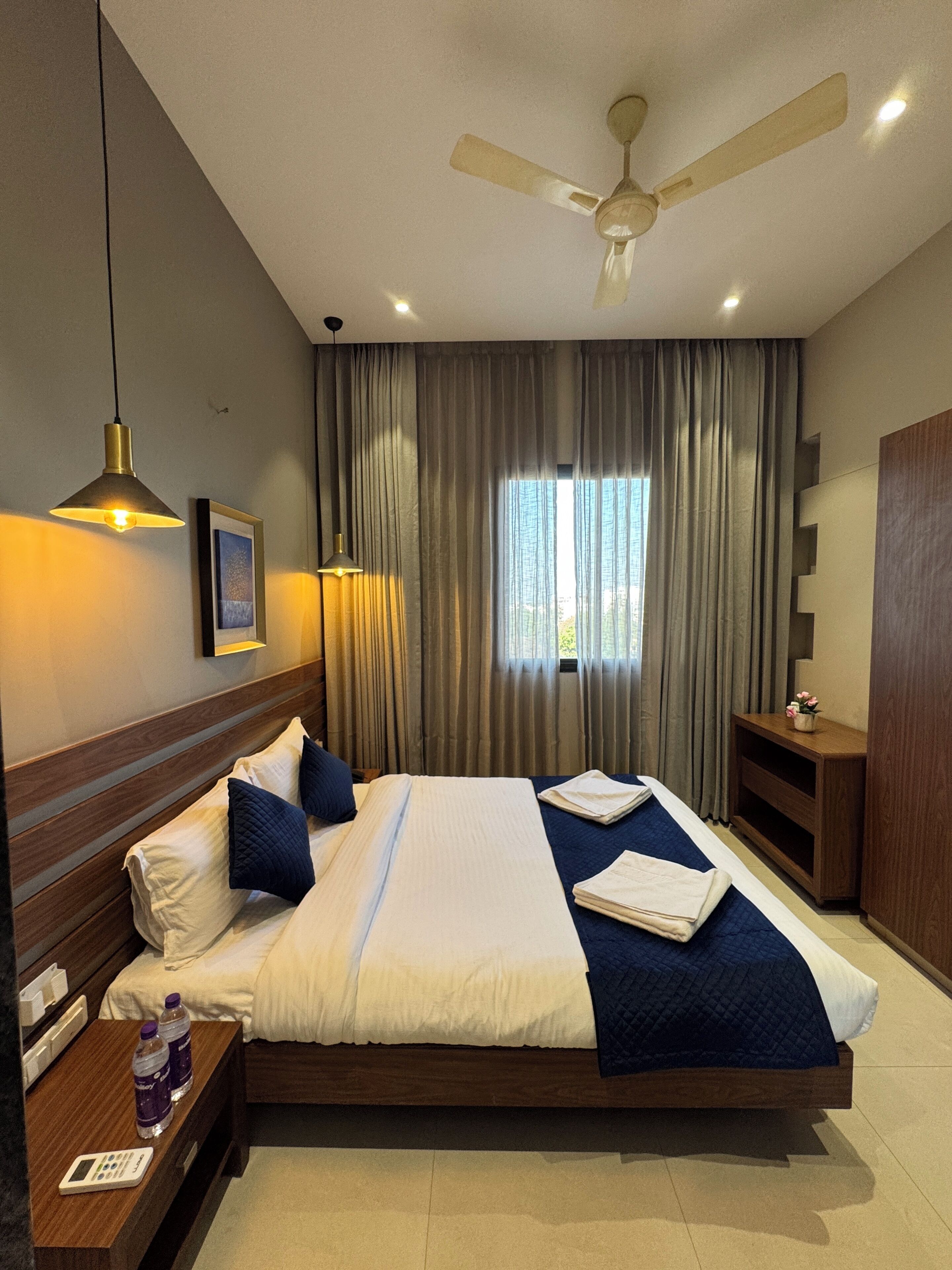 Una habitación de hotel con cama, mesitas de noche, ventilador de techo y ventana con cortinas.