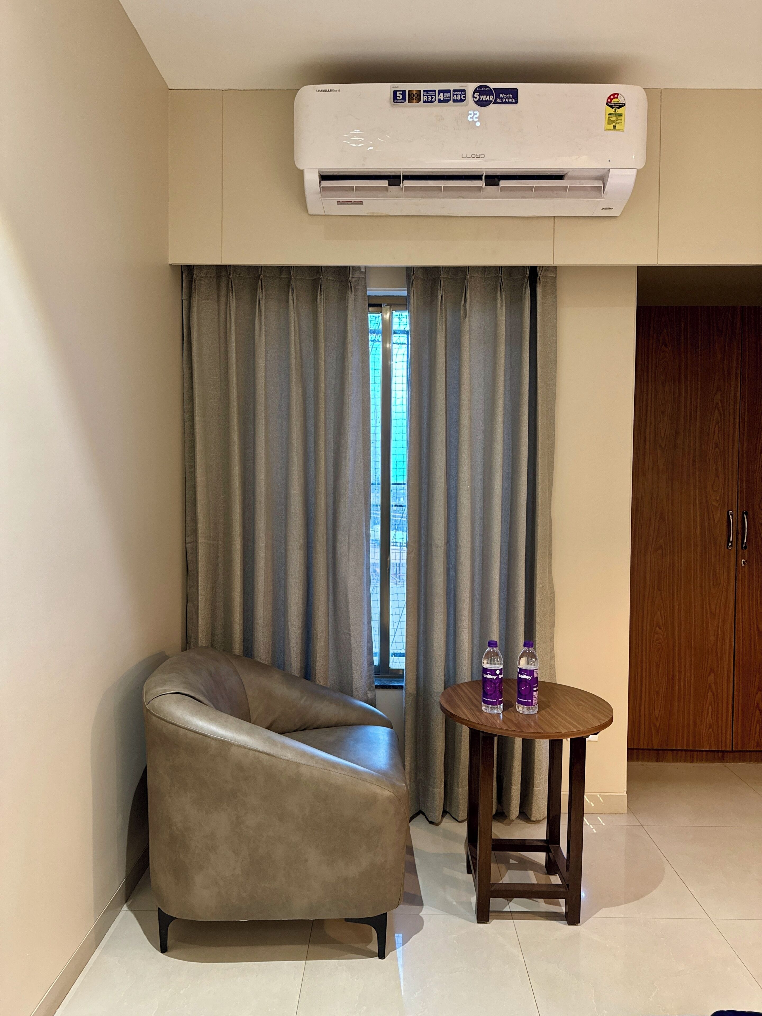 A room with a beige sofa, a small wooden table with a bottle, and an air conditioning unit mounted on the wall.