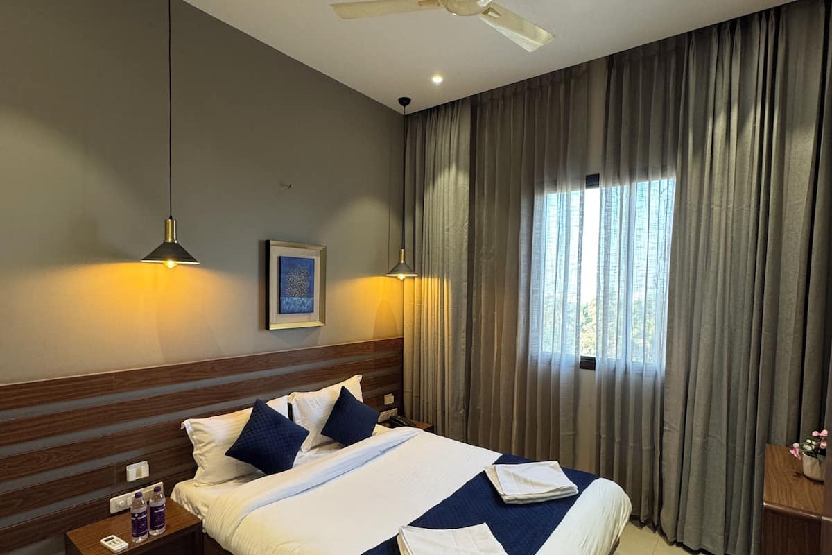 Camera d'albergo con un letto grande, una ventola a soffitto, un comodino con un telefono e bottiglie d'acqua, e una finestra con tende.