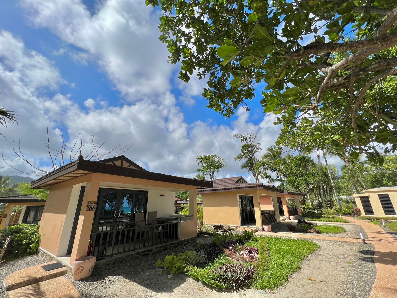 A two-story building with a flat roof and a balcony. There is a sign indicating the location as 'Beach Casita 109'. The building is surrounded by greenery and has a clear sky with clouds.