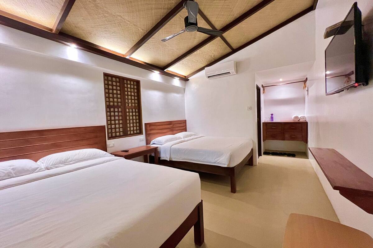 Habitación de hotel con dos camas, cabecero de madera, ventilador de techo, unidad de aire acondicionado, televisor montado en la pared y un pequeño banco de madera.