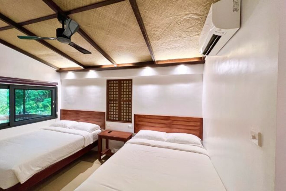 A hotel room with two single beds, a wooden headboard, a window with a view of greenery, a ceiling fan, and an air conditioning unit.