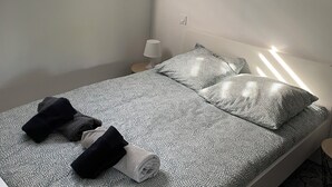 Siististi pedattu sänky kuviollisilla vuodevaatteilla, yöpöydällä oleva lamppu ja ikkunassa olevat sälekaihtimet.