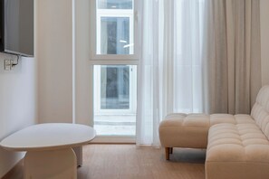 A modern living room with a beige sofa, a round white table, and a large window with sheer curtains.