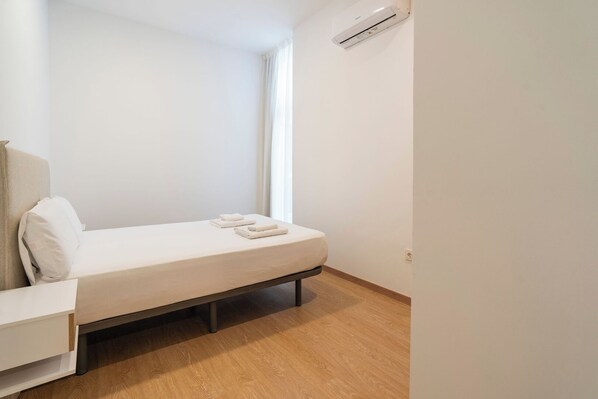 A small, minimalist bedroom with a single bed, a bedside table, and a wall-mounted air conditioner.