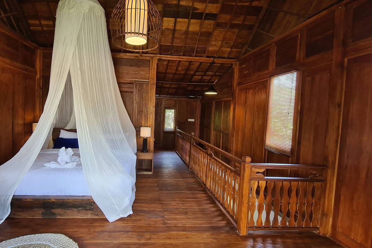 Una habitación de madera con una cama bajo un dosel, una mesita de noche con una lámpara y una barandilla de madera a lo largo del pasillo.