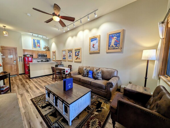A living room with a brown leather sofa, a wooden coffee table, and a patterned rug. There is a kitchen area with a red refrigerator and a microwave. The room has a ceiling fan and several framed posters on the wall. - Arapahoe Lodge 8102 OFB (Keystone)