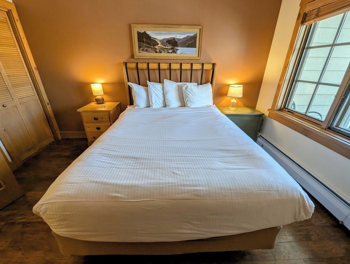 Una cama bien hecha con cabecera, dos mesitas de noche con lámparas, un cuadro sobre la cama y una ventana a la derecha.