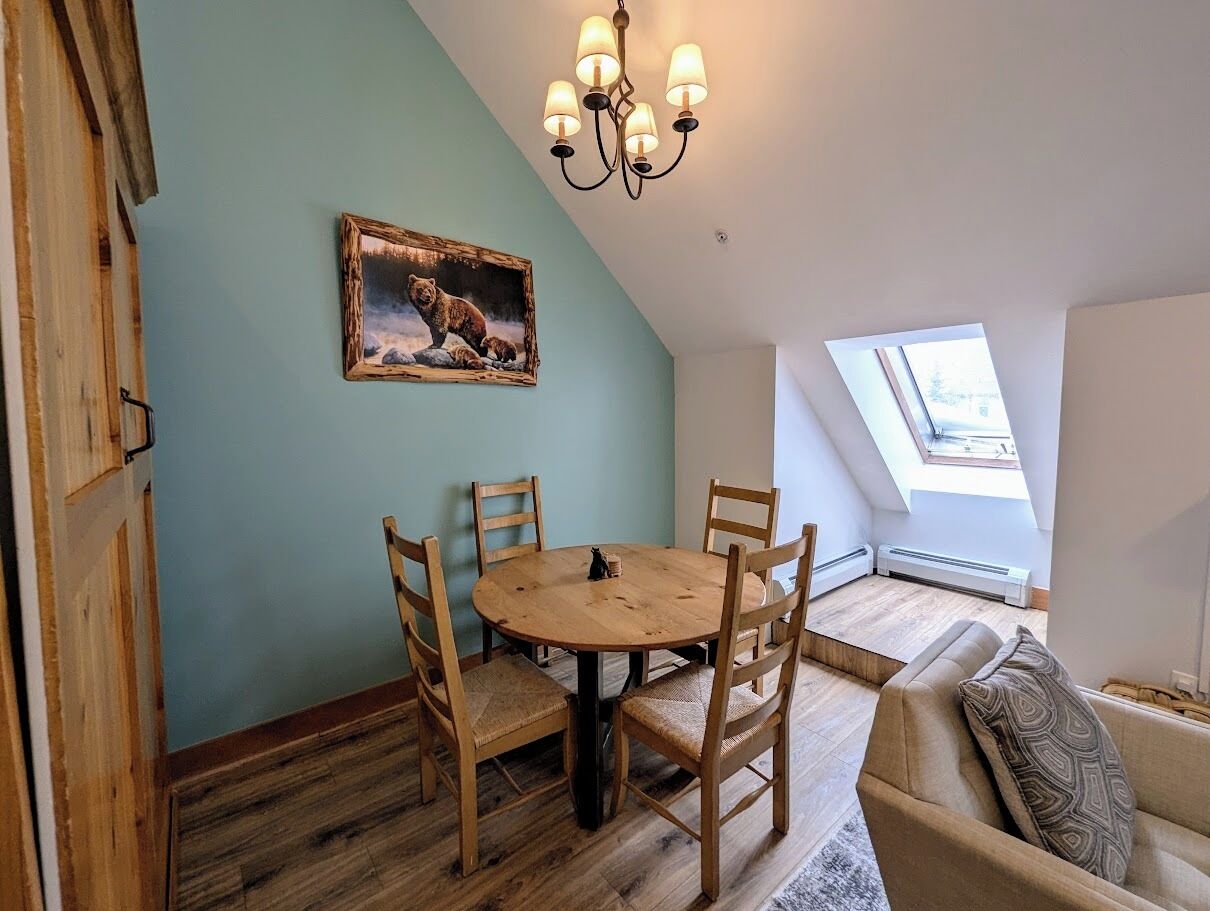 A dining area with a round wooden table and four chairs, a chandelier, a painting on the wall, and a skylight.