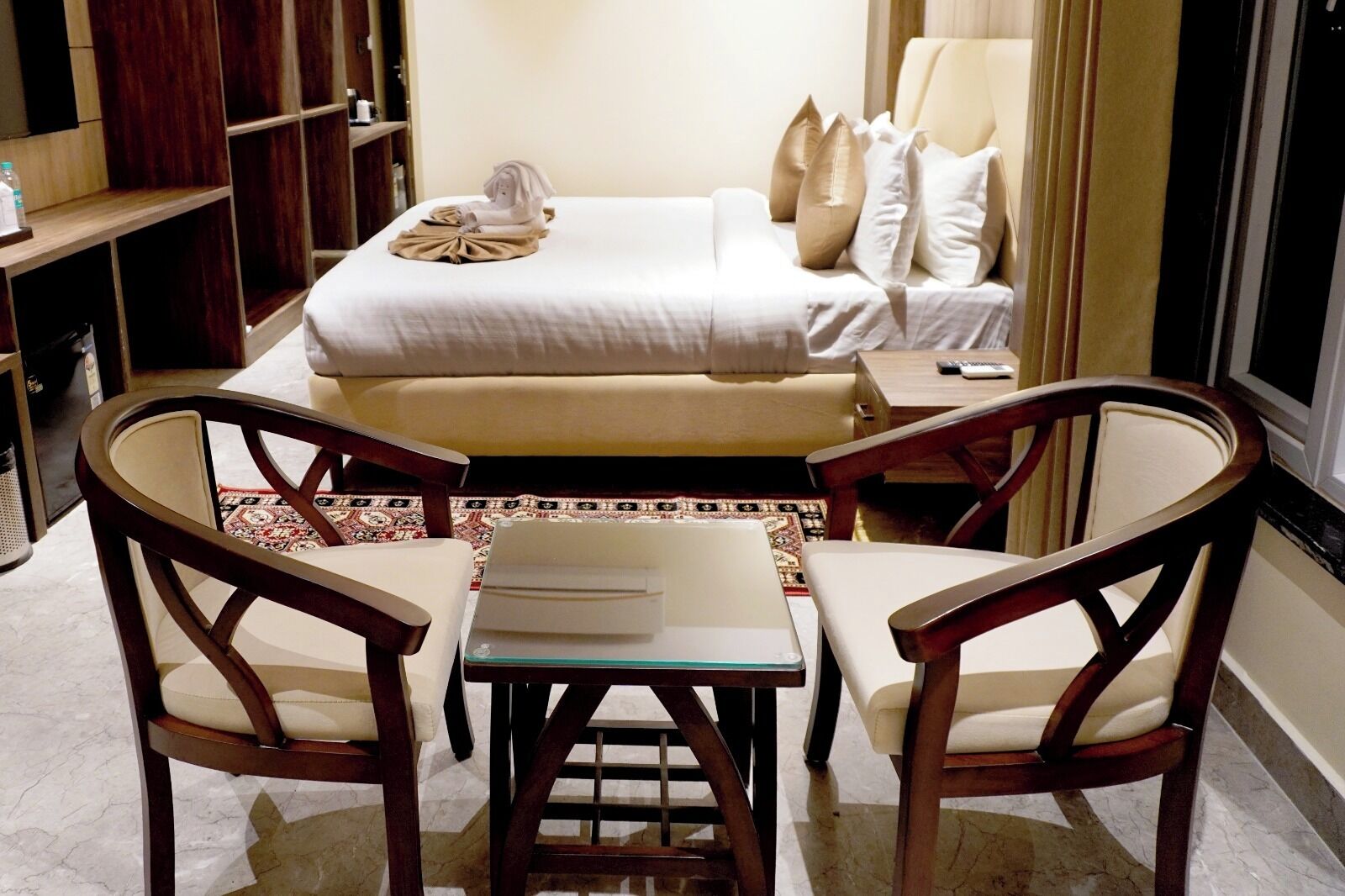A hotel room with a bed, two chairs, a small table, and a bookshelf.