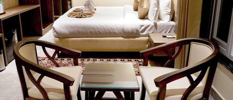 A hotel room with a bed, two chairs, a small table, and a bookshelf.