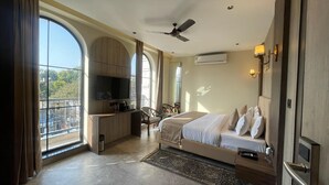 A modern hotel room with a large bed, a desk with a flat-screen TV, a ceiling fan, and a view of the city through large windows. - HOTEL HRISHIKESH HIGH (Rishikesh)