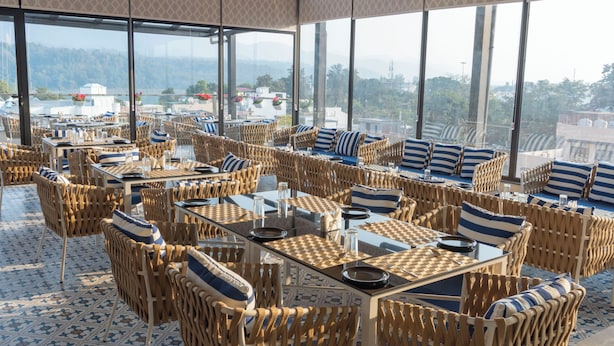 A spacious outdoor dining area with wicker furniture and checkered tablecloths, large windows offering a view of the surrounding landscape. - HOTEL HRISHIKESH HIGH (Rishikesh)