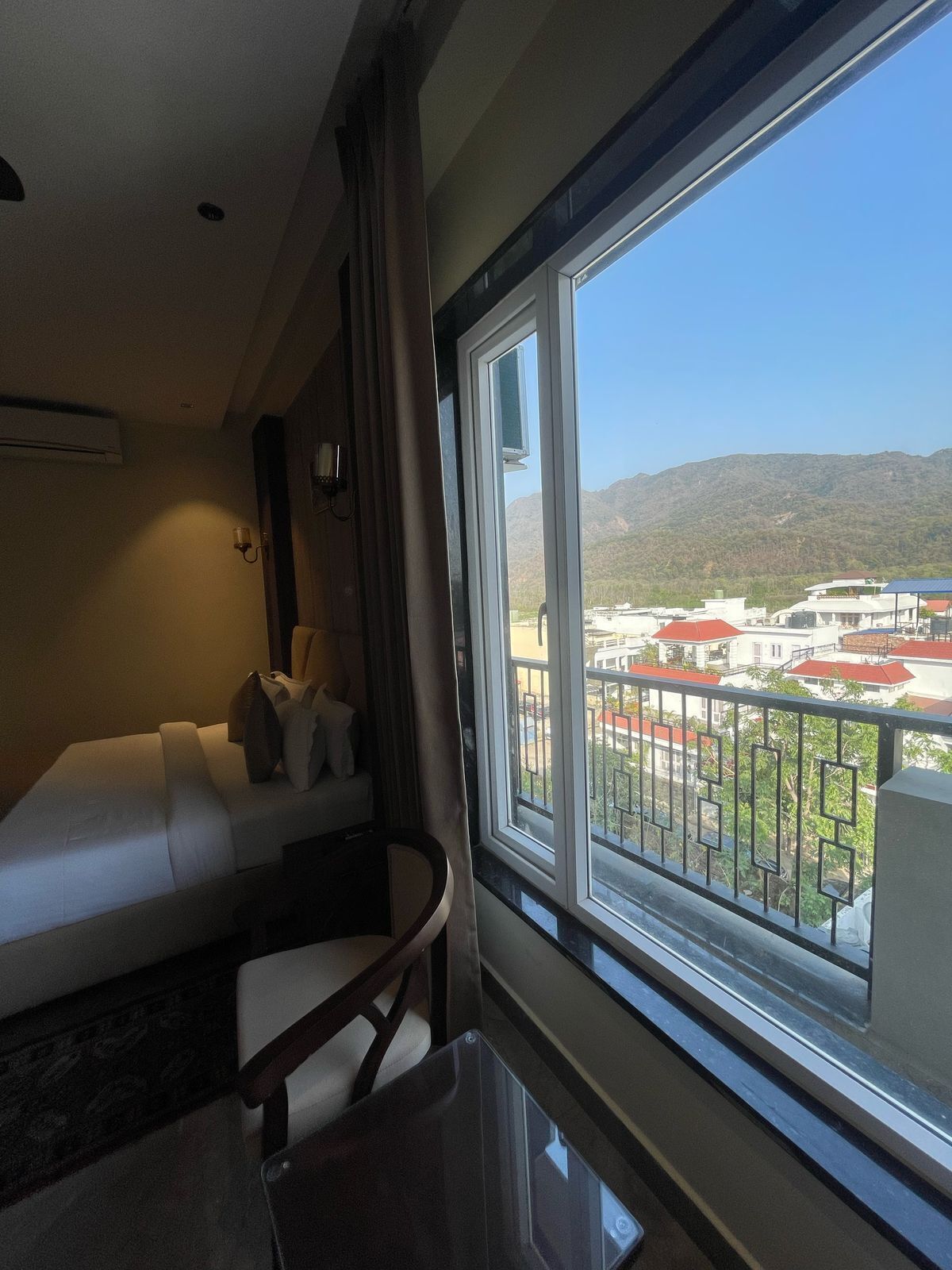 A hotel room with a bed, a small table, a chair, a window with a view of buildings and mountains.