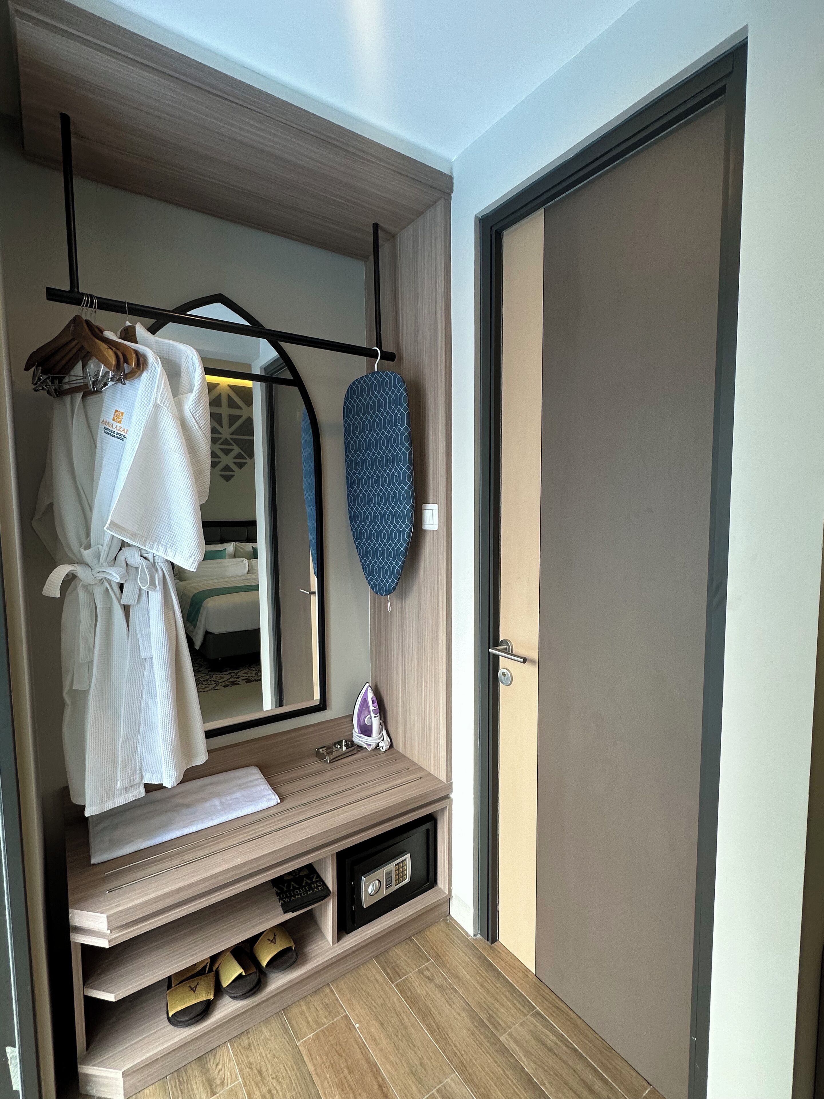 Une chambre d’hôtel équipée d’une coiffeuse en bois, d’un miroir, d’un peignoir suspendu, d’une planche à repasser et d’un coffre-fort.