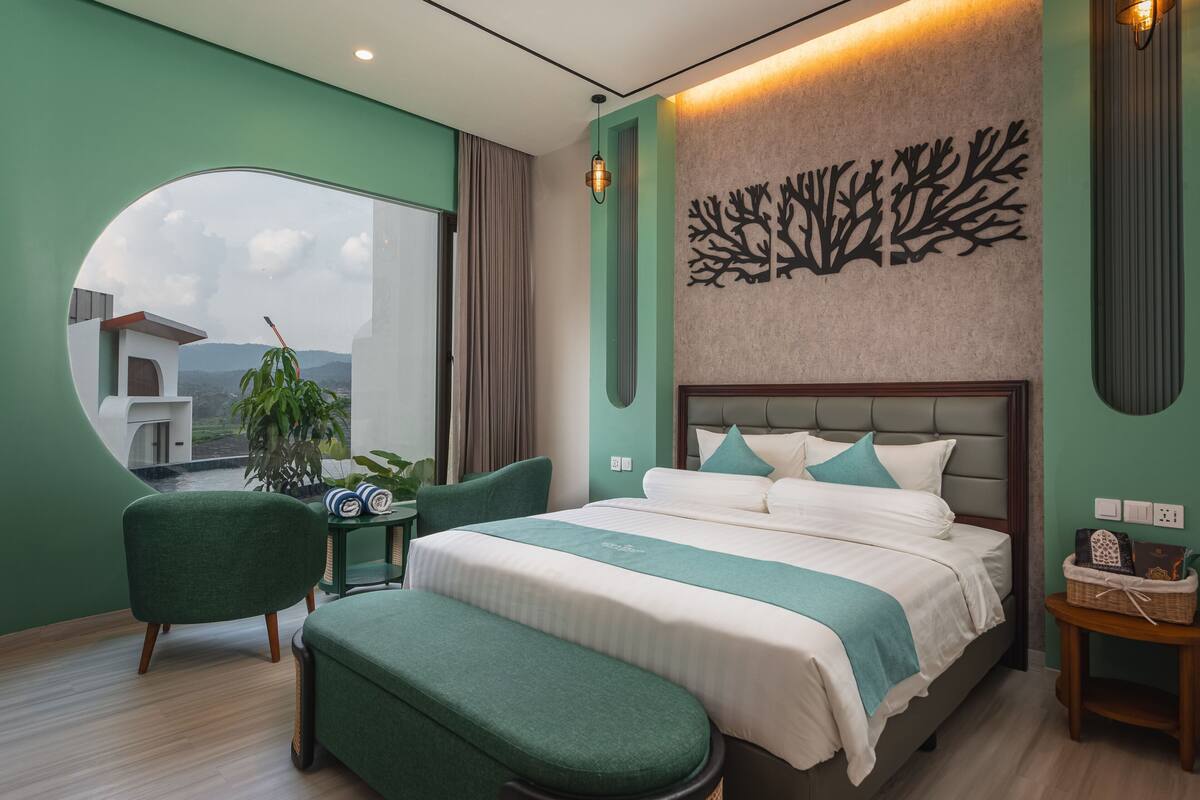 Une chambre d’hôtel moderne avec un grand lit, un fauteuil vert, une petite table et une vue sur l’extérieur.